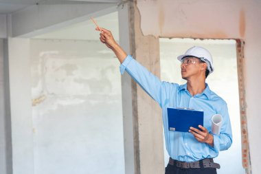 İnşaat mühendisi planı tutuyor. İnşaat alanında çalışan işçilerden mavi gömlekli, güvenlik önleyici beyaz şapka giyiyor. Mimarlık mühendisi, inşaat mühendisi, emlak konseptine bakıyor.