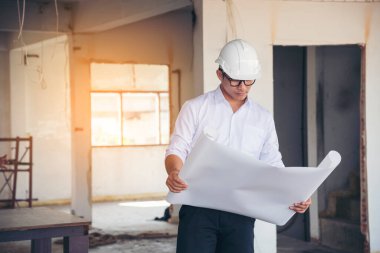 İnşaat mühendisi planı tutuyor. İnşaat alanında çalışan işçilerden mavi gömlekli, güvenlik önleyici beyaz şapka giyiyor. Mimarlık mühendisi, inşaat mühendisi, emlak konseptine bakıyor.