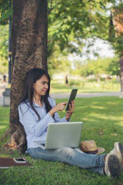 Yeşil bahçe parkında oturan Asyalı kadın telefon kullanarak bilgisayarlı akıllı telefondan müzik dinliyor. Mutluluk Kadını gülümsüyor, gülüyor, mutlu dişlek gülümsüyor. Ev bilgisayarında serbest çalışıyor..