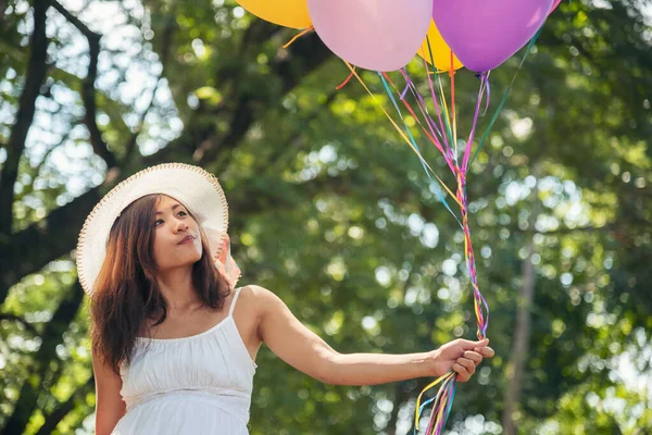 Cheerful beauty woman holding balloons relax sitting under big tree in green park with happiness. Woman Hands holding vibrant air balloons play on birthday party happy time summer on sunshine outdoor