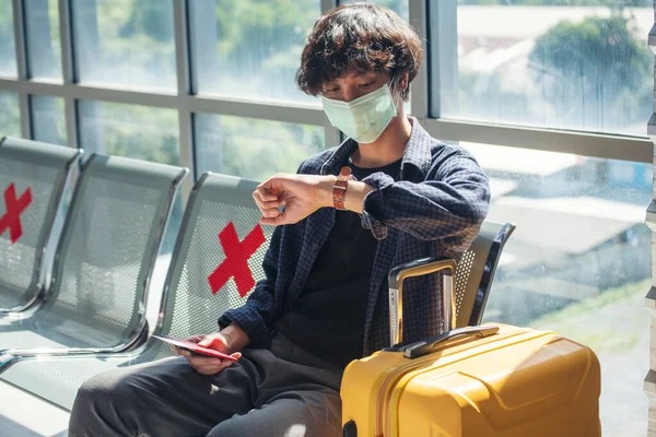 Asian young man traveller new normal wearing face mask sitting Social distancing holding smartphone look at wristwatch in Airplane lounge. New normal Male plane travel waiting passenger boarding time