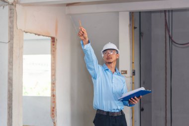 İnşaat mühendisi planı tutuyor. İnşaat alanında çalışan işçilerden mavi gömlekli, güvenlik önleyici beyaz şapka giyiyor. Mimarlık mühendisi, inşaat mühendisi, emlak konseptine bakıyor.