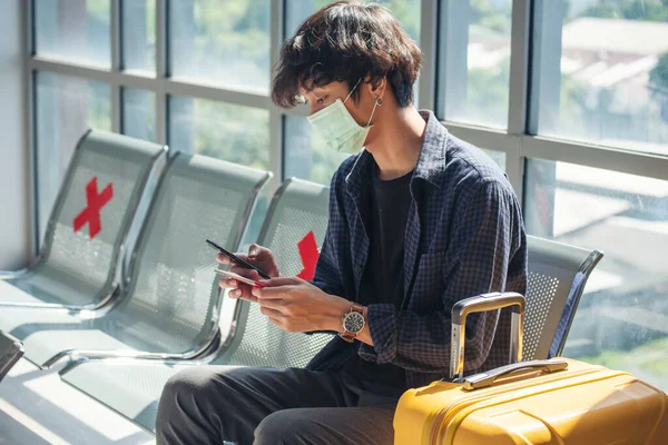 Asian young man traveller new normal wearing face mask sitting Social distancing holding smartphone in Airplane lounge. New normal Male travel pandemic passenger by plane. Coronavirus tourism trip