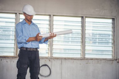 İnşaat mühendisi planı tutuyor. İnşaat alanında çalışan işçilerden mavi gömlekli, güvenlik önleyici beyaz şapka giyiyor. Mimarlık mühendisi, inşaat mühendisi, emlak konseptine bakıyor.