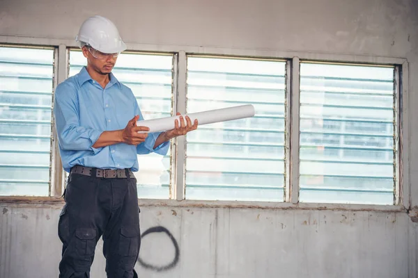 İnşaat mühendisi planı tutuyor. İnşaat alanında çalışan işçilerden mavi gömlekli, güvenlik önleyici beyaz şapka giyiyor. Mimarlık mühendisi, inşaat mühendisi, emlak konseptine bakıyor.