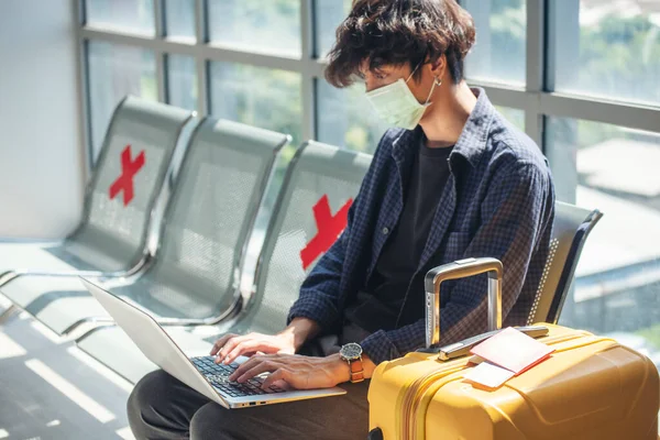 Asian young man traveller new normal typing laptop keyboard wear face mask Social distancing in Airplane lounge. New normal Male travel pandemic passenger by plane. New normal tourist work anywhere