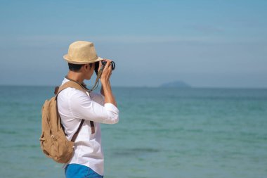 Man traveler using digital camera on summer beach blue sky take a photo. Asian Photographer man journey relax on the beach in summertime. Man backpack shooting photo asia destination holiday trip