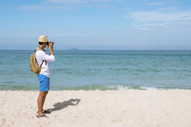 Man traveler using digital camera on summer beach blue sky take a photo. Asian Photographer man journey relax on the beach in summertime. Man backpack shooting photo asia destination holiday trip
