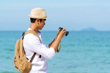 Man traveler using digital camera on summer beach blue sky take a photo. Asian Photographer man journey relax on the beach in summertime. Man backpack shooting photo asia destination holiday trip