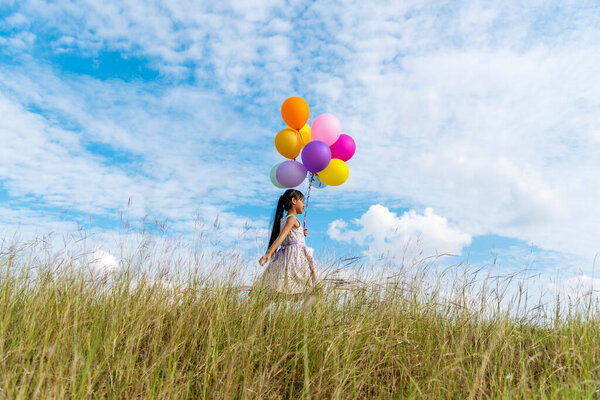 Cheerful cute girl holding balloons running on green meadow white cloud and blue sky with happiness. Hands holding vibrant air balloons play on birthday party happy times summer on sunlight outdoor