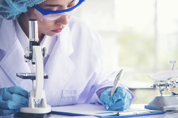 Asian Woman scientist hand write note research in medical lab look at science microscope medical test. Researcher biology chemistry. Asian Female technician laboratory using microscope blood test