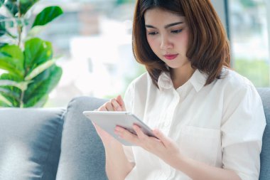 İnternetten dijital tablet alışverişi yapan Asyalı bir kadın arıyor, mesaj yolluyor, internet teknolojisi yaşam tarzında gülümsüyor. Asyalı kadın akıllı tablete dokunuyor mobil uygulama ev ofisini kullanarak ellere odaklanıyor.