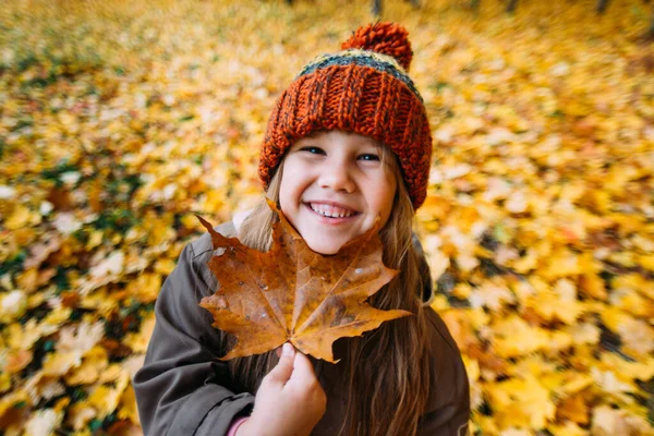 Ein kleines Mädchen steht in einem herbstlichen Park mit einem Ahornblatt und lächelt. — Stockfoto