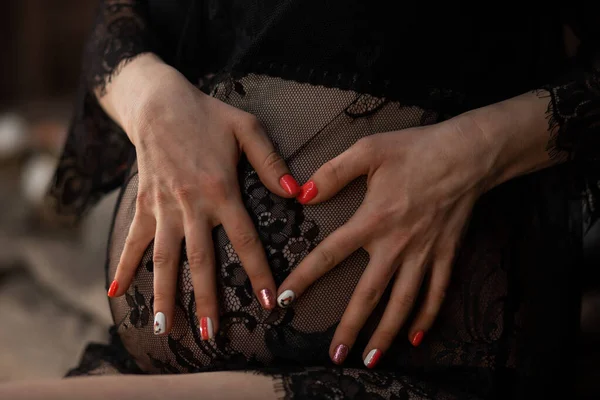 Bir kızın hamile karnının arka planında parmaklarından yapılmış bir kalp. Siyah dantelli elbiseyle