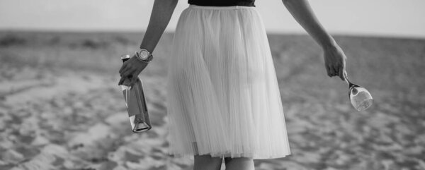 A girl walks along the sand of the beach in a white skirt with a bottle and a glass of wine. Black and white photo