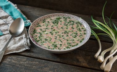 Ahşap bir masada bir tabak okroshka. Havluda kaşık ve yeşil soğan.