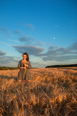 A girl with a bouquet of wheat ears stands in a wheat field at sunset