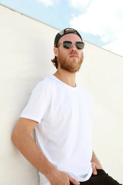 Portré szakállas férfi pózol szabadtéri visel napszemüveg és fehér póló. — Stock Fotó