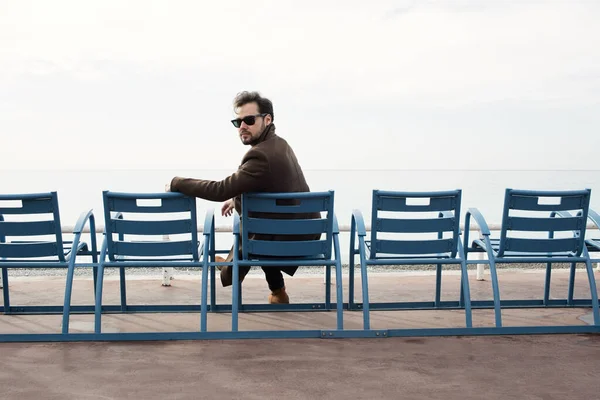 Szakállas férfi kabátban ül a padon a tenger partja mellett.. — Stock Fotó
