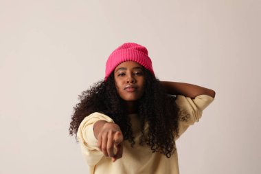 Kendine güvenen siyah kadın beyaz duvarın üzerinde duruyor ve parmağını kameraya doğrultuyor..