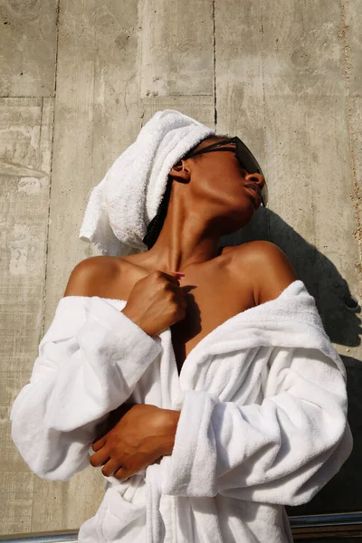 Jolie jeune femme enveloppée dans une serviette blanche posant sur un mur gris. Vertical. — Photo