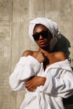 Beyaz havluya sarılmış, gri duvarda poz veren çekici genç bir kadın. Dikey.