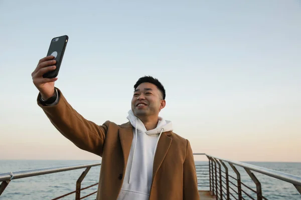 Knappe jonge Aziatische man met behulp van zijn smartphone outdoor het maken van een selfie portret. — Stockfoto