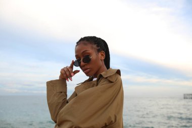 Güneş gözlüğü ve yağmurluk takan, dışarıda poz veren muhteşem siyahi bir kadın..