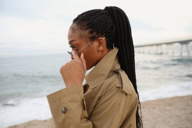 Siyahi bir kadının güneş gözlüğü ve yağmurluk takıp açık havada poz verirken çekilmiş fotoğrafı..