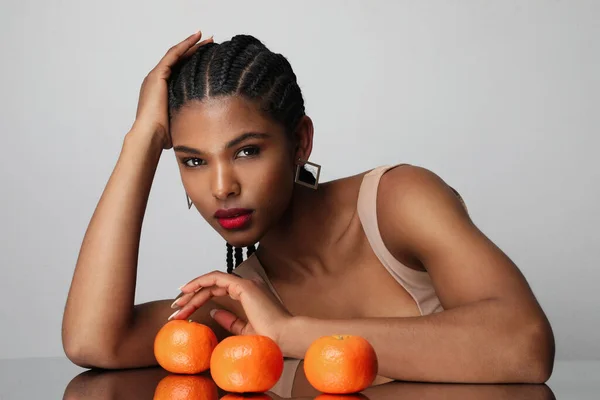 African American woman with long dark braids and orange fruits poses indoor. High quality photo.