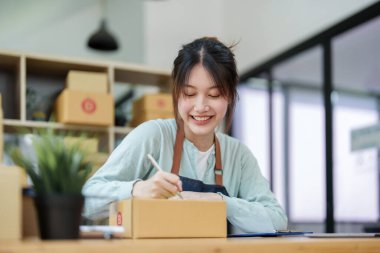 Küçük bir girişimin portresi, ve KME sahibi, Asyalı bir kadın girişimci, müşterinin iç kutusuna koymadan önce ürünü organize etmek için bir not defterine bilgi yazıyor.