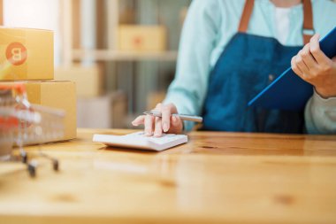 Portrait of a small startup, SME owner, Asian female entrepreneur, checks online orders and uses a price calculator to calculate product prices and packs products into boxes to customers