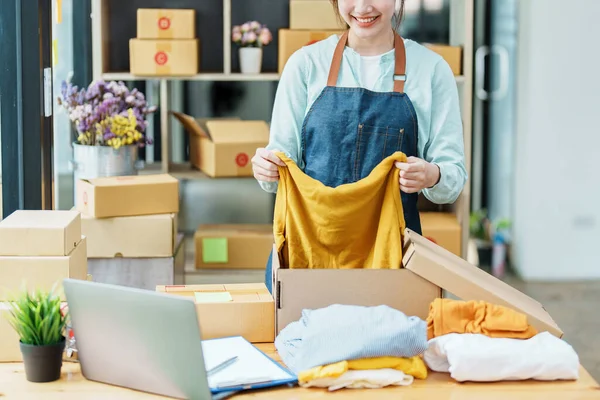 Portrait of a small startup Asian female entrepreneur SME owner picking up a yellow shirt before packing it in an inner box with a customer. Online Business Ideas and Freelance — Φωτογραφία Αρχείου