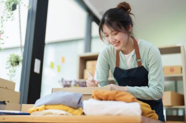 Küçük bir girişimin portresi, ve KME sahibi, Asyalı bir kadın girişimci, müşterinin iç kutusuna koymadan önce ürünü organize etmek için bir not defterine bilgi yazıyor..