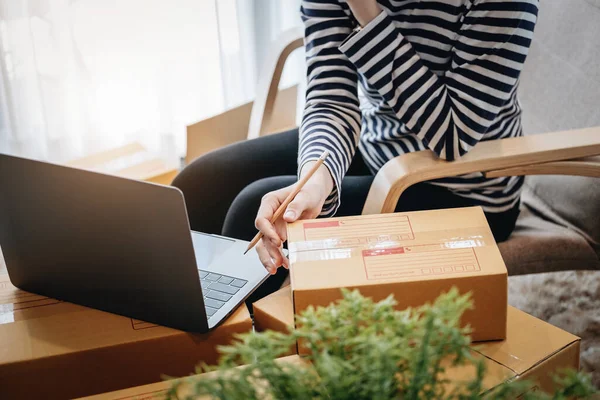 Online merchandising business idea, beautiful girl holding a parcel delivery box and using a computer to input Track And Trace parcel numbers to customers