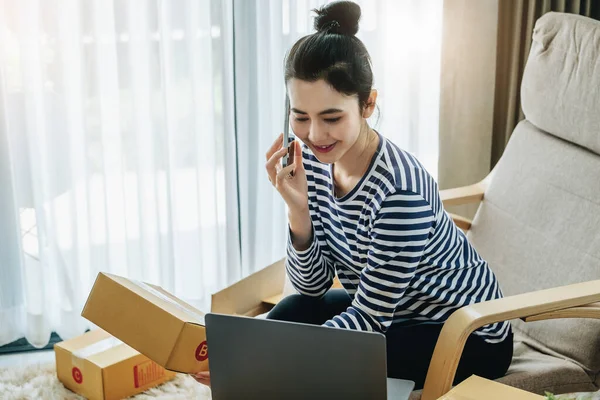 Online merchandising business idea, beautiful girl talking on the phone and holding a parcel box ready to use a computer to enter the Track And Trace parcel number to the customer