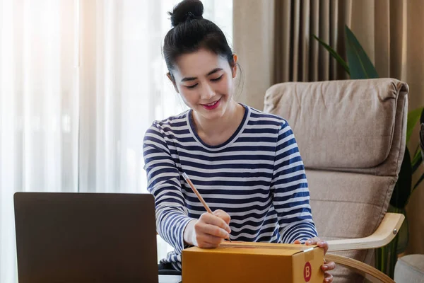 Online selling business idea, beautiful girl writing customer address in front of parcel delivery box and using computer to enter tracking and trace number of parcels for customers