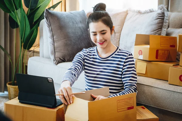 Online merchandising business idea, a beautiful girl is packing the products in the parcel box from the order information that the customer ordered on the tablet computer