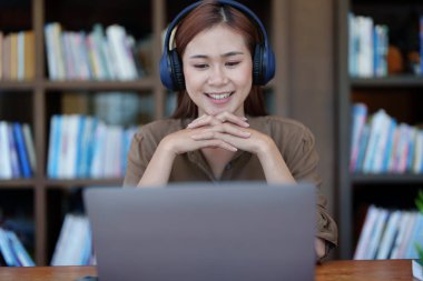 Kulaklık takan, gülümseyen Asyalı genç bir kızın portresi ve bir kütüphanede online video konferansı için bilgisayar kullanımı..