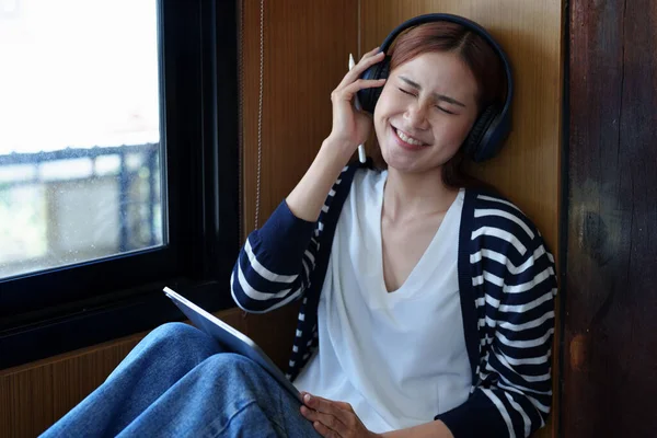 一个年轻的亚洲女人的画像 她面带微笑 用平板电脑 戴着耳机 高高兴兴地坐在那里休息 — 图库照片