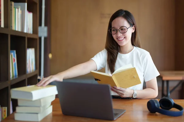 Portrait of a teenage Asian woman using a computer and notebook to study online via video conferencing on a wooden desk in library.