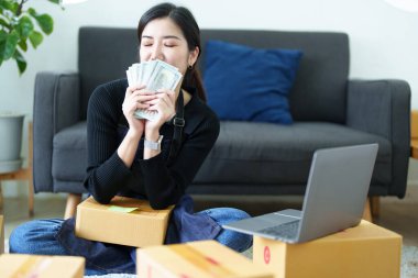 Çevrimiçi pazarlama paketi ürünleri ve KME dağıtım kavramının neşeli başarısıyla dizüstü bilgisayarla gülümseyen ve elinde para tutan bağımsız Asyalı kadının küçük işletme girişimciliğini başlatan.