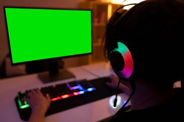 Blurred green computer screen, selective focus on teenage who is using computer in a dark room. Teenage wearing a headset with a mic, dark room with warm led lights.