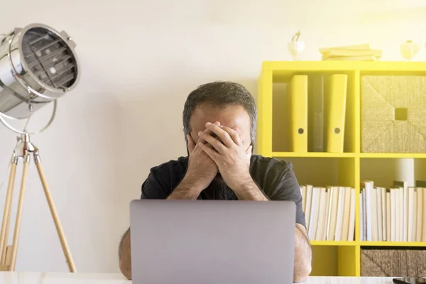 Depressed man, front view of young bearded depressed man or stressed or sad businessman at the office. Sitting at the table, covered his face with hands. Working on laptop and got bad news.