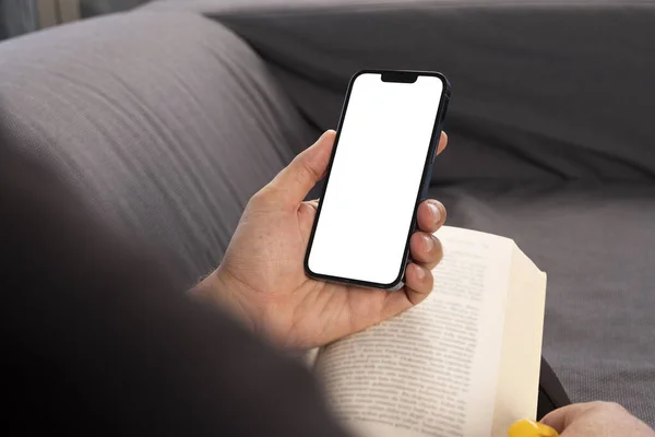 Holding mobile phone, over shoulder view of man hand holding mobile phone.  Blank white screen for smartphone mock up. Caucasian male relaxing on the couch, reading book and looking to phone display.