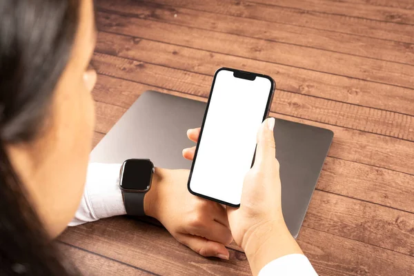Woman holding smartphone,  over shoulder view of woman holding smartphone. Caucasian female wearing smart watch and looking white blank screen of mobile phone for mock up. Sitting on the wooden table.