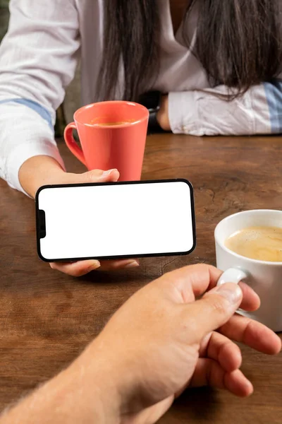 Showing smartphone horizontal,  couple sitting on the wooden table and woman showing smartphone horizontal. Mobile phone mock up concept photo with white blank screen. Drinking hot fresh coffee.