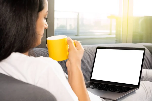 Woman relaxing on the couch, holding modern laptop for mock up and woman relaxing on the couch. Millennial young beautiful lady drinking morning coffee and watching or looking to notebook screen.