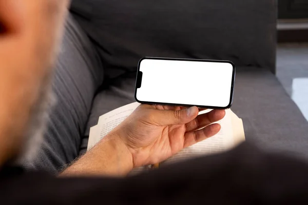 Holding horizontal smartphone, over shoulder view of holding horizontal smartphone. White blank screen for mock up. Man sitting on the couch and watching movie or game or attending online meeting.