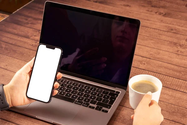 Video chat mock up, woman holding modern phone for video chat mock up. Application recommendation concept idea. Using laptop, drinking coffee and looking white blank screen of smartphone while sitting